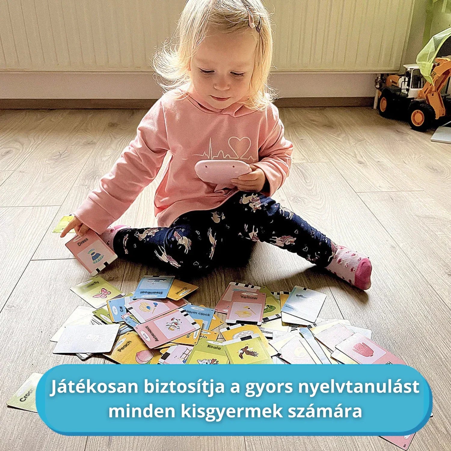 Deklica sedi na tleh in se igra z interaktivno napravo ter številnimi izobraževalnimi karticami za hitro in zabavno učenje jezika.